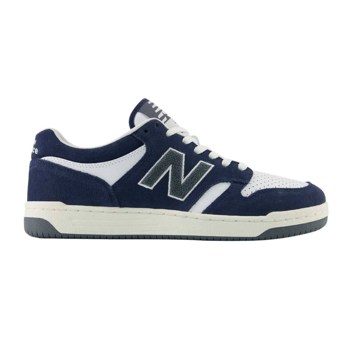 A New Balance sneaker with a white perforated toe, navy blue suede overlays, white laces, a large navy “N” logo outlined in gray, and a white midsole with a dark gray outsole.
