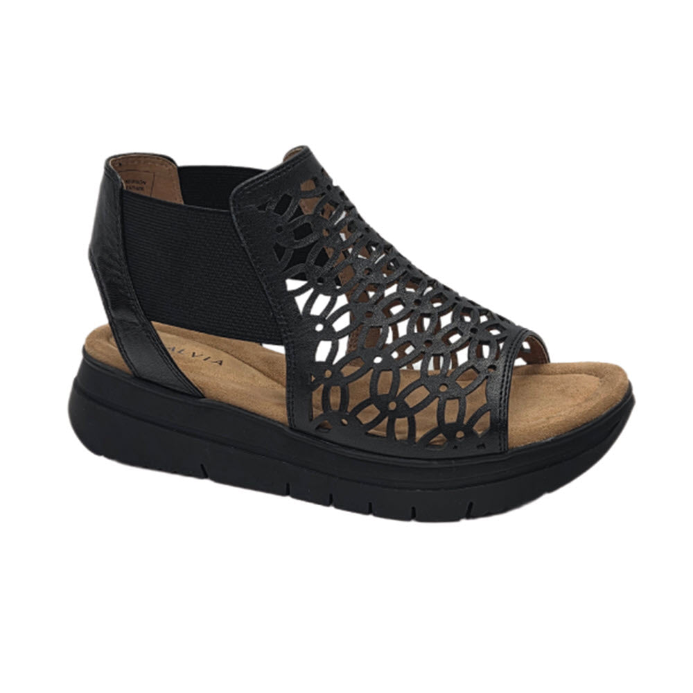 A black open-toe sandal with a thick sole, tan footbed, and a geometric cut-out design on the upper part. The sandal has a closed heel and an elastic strap on the side.