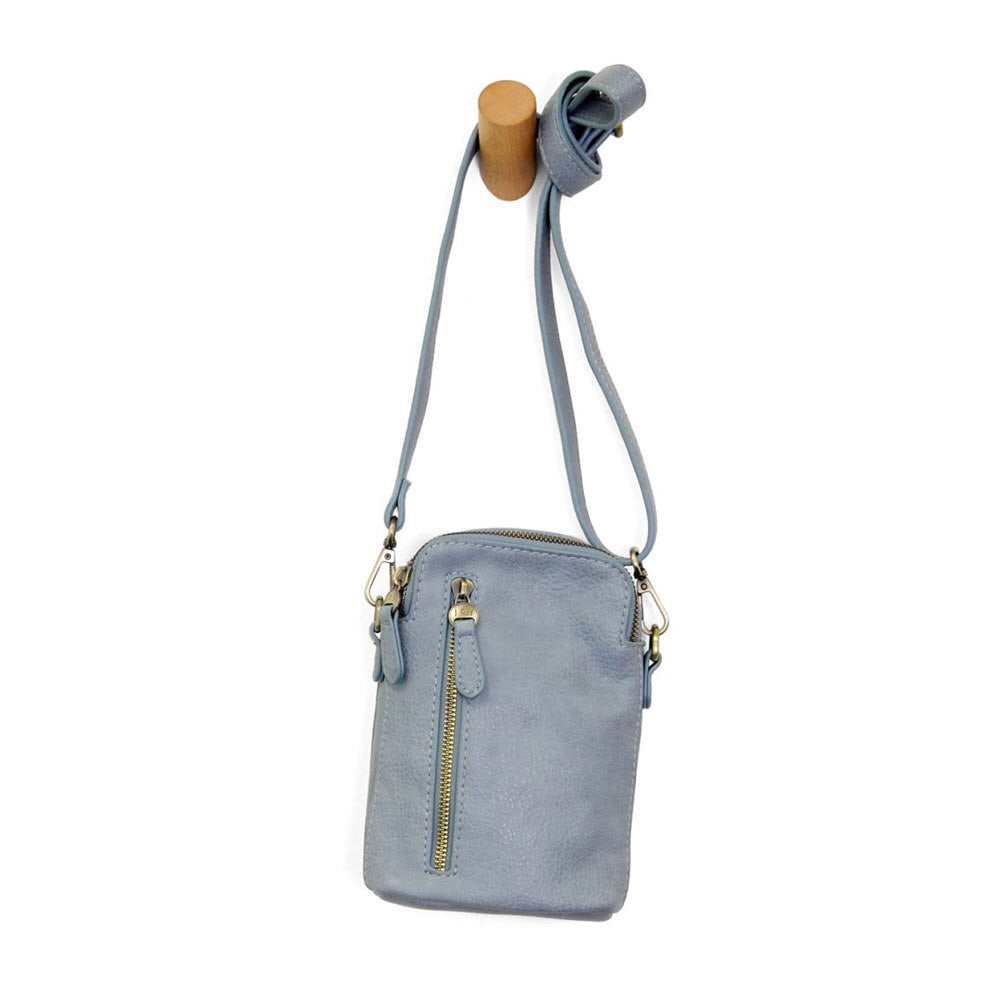 A small light blue leather crossbody bag with a front zipper pocket and adjustable strap, hanging from a wooden peg on a white background.