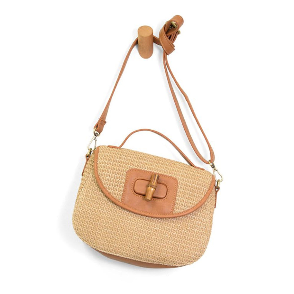 A woven straw crossbody bag with a light brown leather strap, trim, and handle, featuring a wooden clasp on the front, hanging on a wooden wall hook.