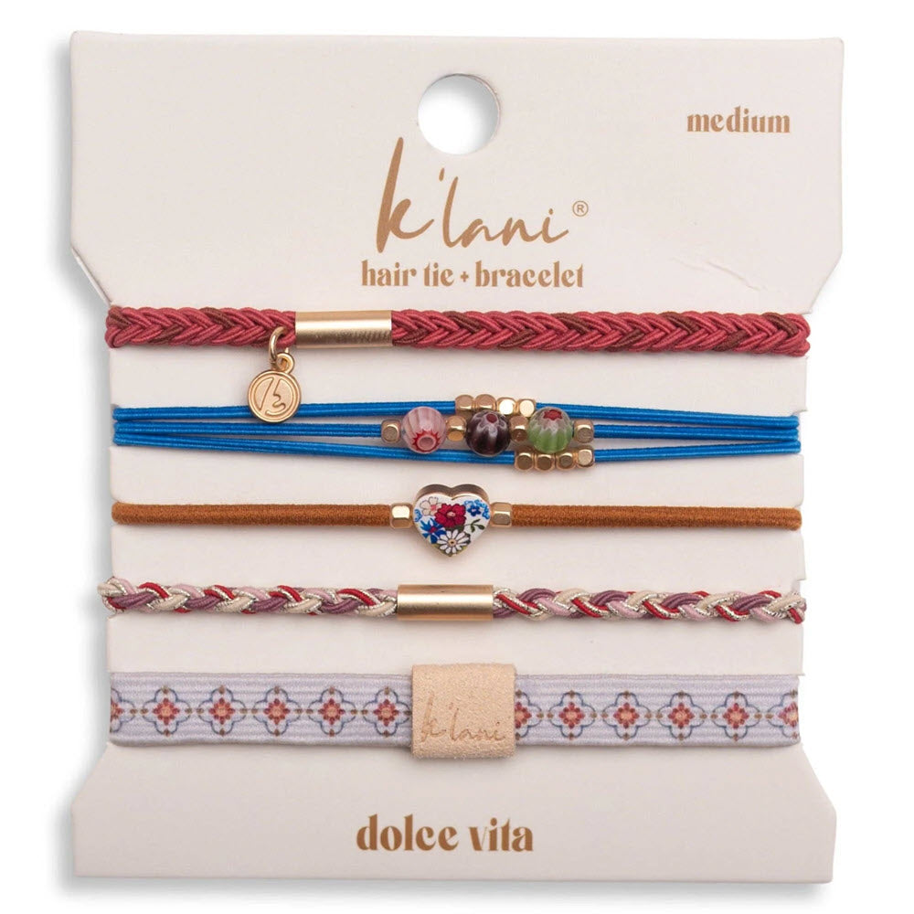 Five decorative hair ties and bracelets in various colors and styles, including braids, beads, and charms, displayed on a white card labeled “k’lani,” “medium,” and “dolce vita.”.