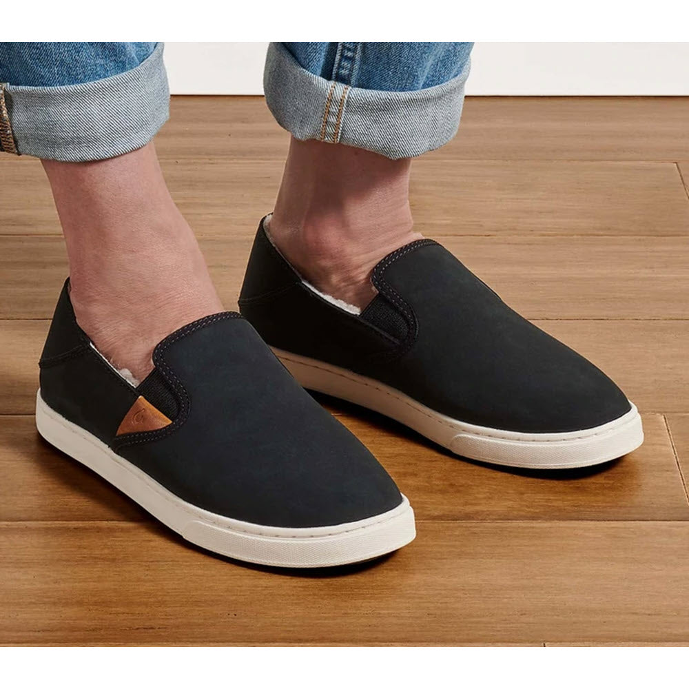 A person wearing Olukai Pehuea Heu Lava Rock/Lava Rock - Womens slip-on sneakers with waterproof nubuck leather and white soles stands on a wooden floor.