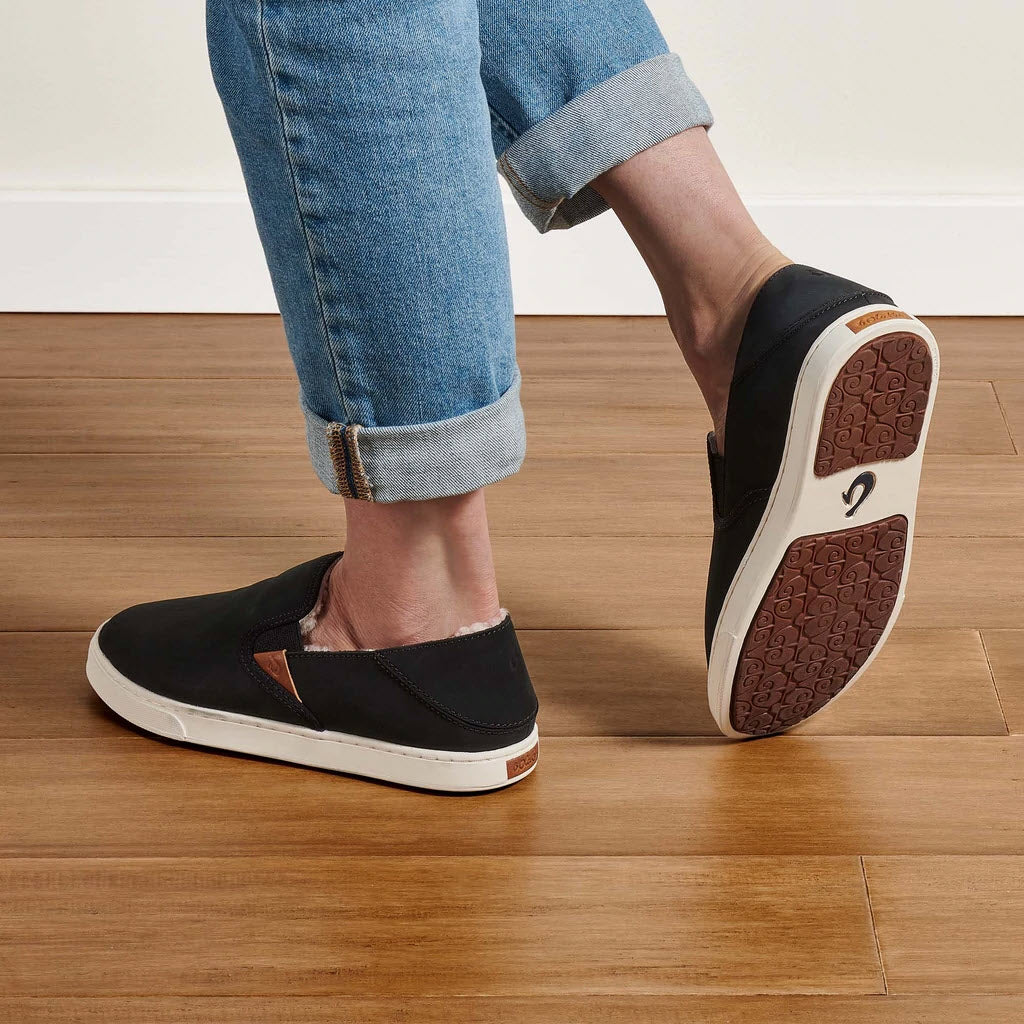 Person wearing Olukai Pehuea Heu Lava Rock/Lava Rock - Womens slip-on sneakers with durable sneaker outsole and white soles, standing on a wooden floor.