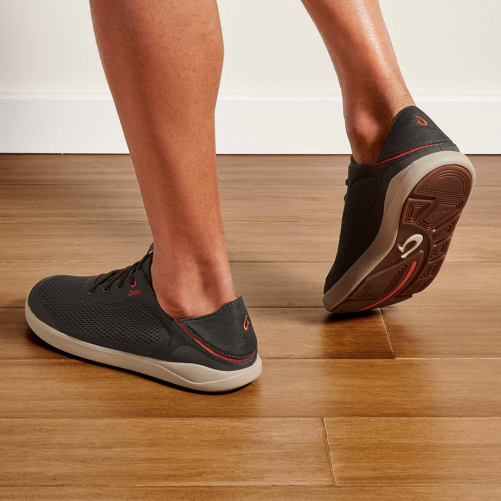 A person wearing Olukai Moku Pae Island Salt/Koi athletic shoes taking a step on a wooden floor.
