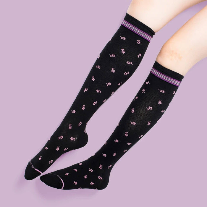 A pair of legs wearing black Dr. Motion compression socks with a floral pattern, against a purple background.