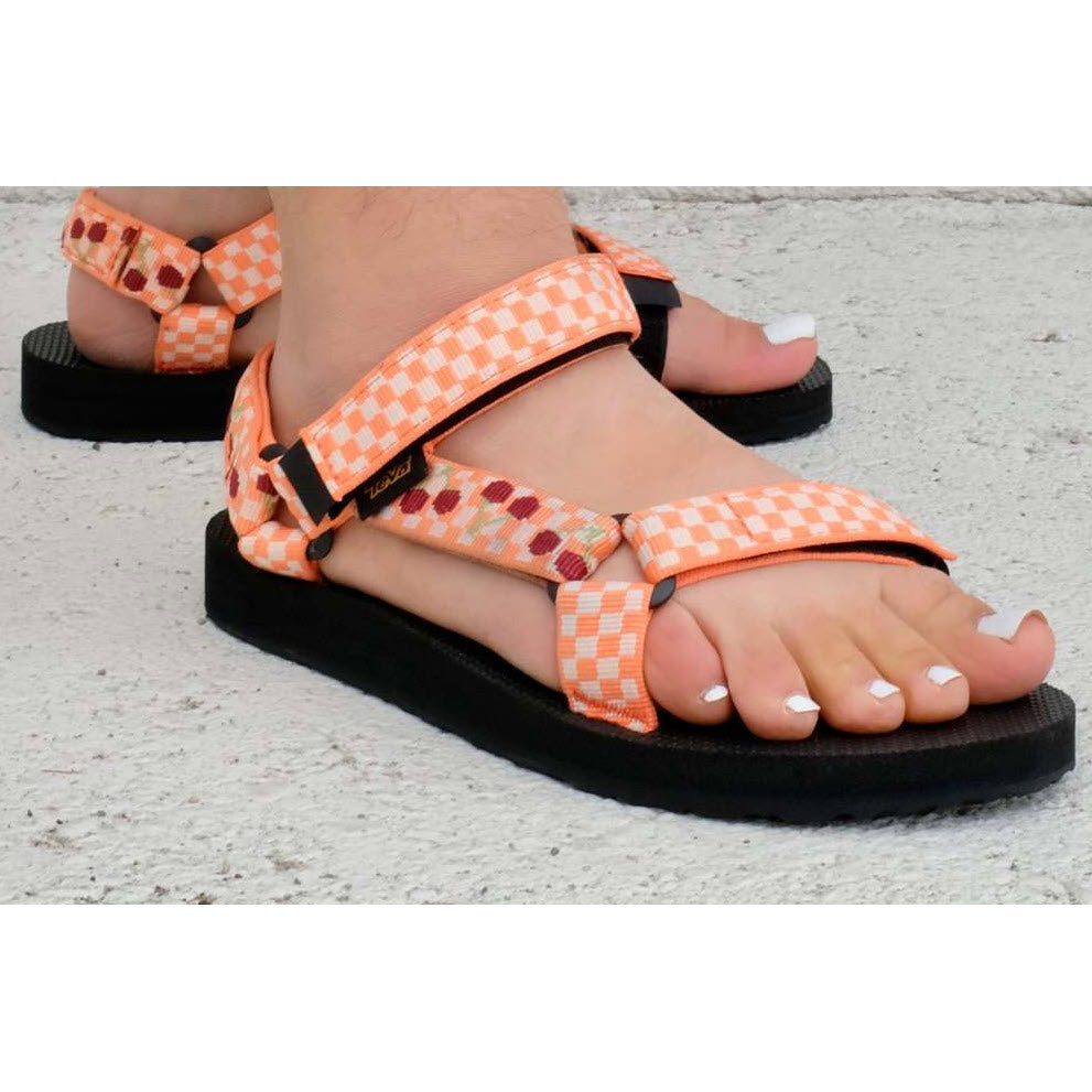 A close-up of a person&#39;s feet wearing Teva Original Universal Picnic Cherries - Kids water sandals with recycled plastic straps on a concrete surface.