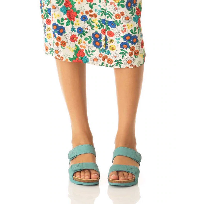A person standing in teal Dansko Maddy Lagoon Milled Nubuck slide sandals with leather uppers and a floral skirt.