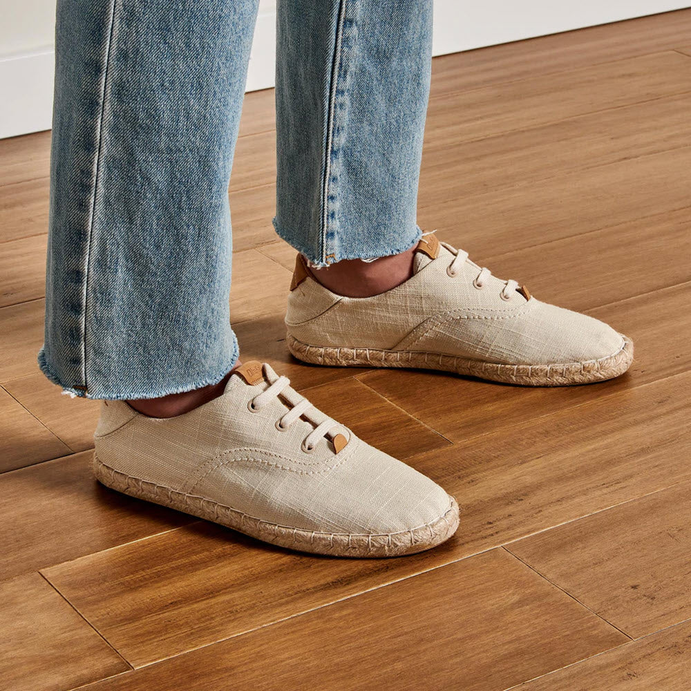 Person standing on a wooden floor wearing beige Olukai KAULA PA&#39;A LI TAPA espadrille shoes and blue denim jeans.