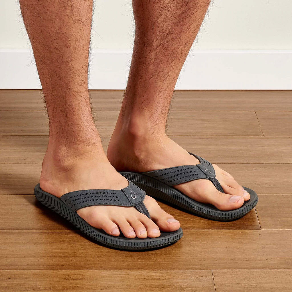 A person wearing gray Olukai Ulele Dark Shadow - Mens water-friendly sandals on a wooden floor.