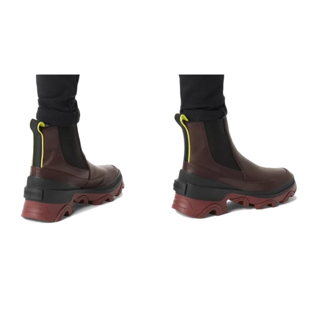 A pair of SOREL BREX BOOT CHELSEA NEW CINDER - WOMENS with a high-traction rubber sole, worn by someone with black pants against a white background.