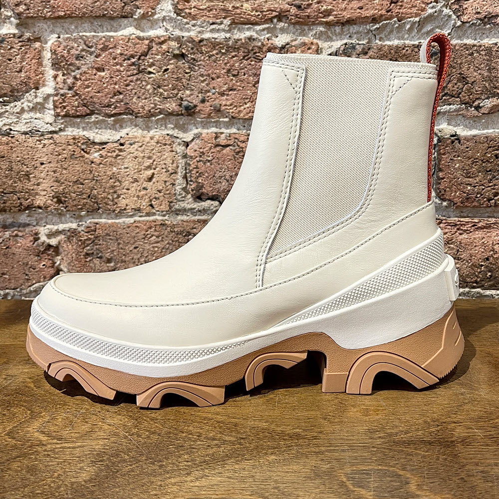 A beige Sorel Brex Boot Chelsea Natural - Womens with elastic side panels and a red pull-tab against a brick wall background.