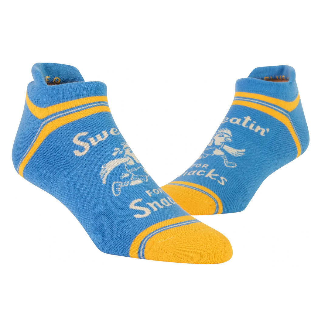 A pair of Blue Q ankle workout socks with yellow accents and a humorous phrase "sweatin' for snacks" printed on them.