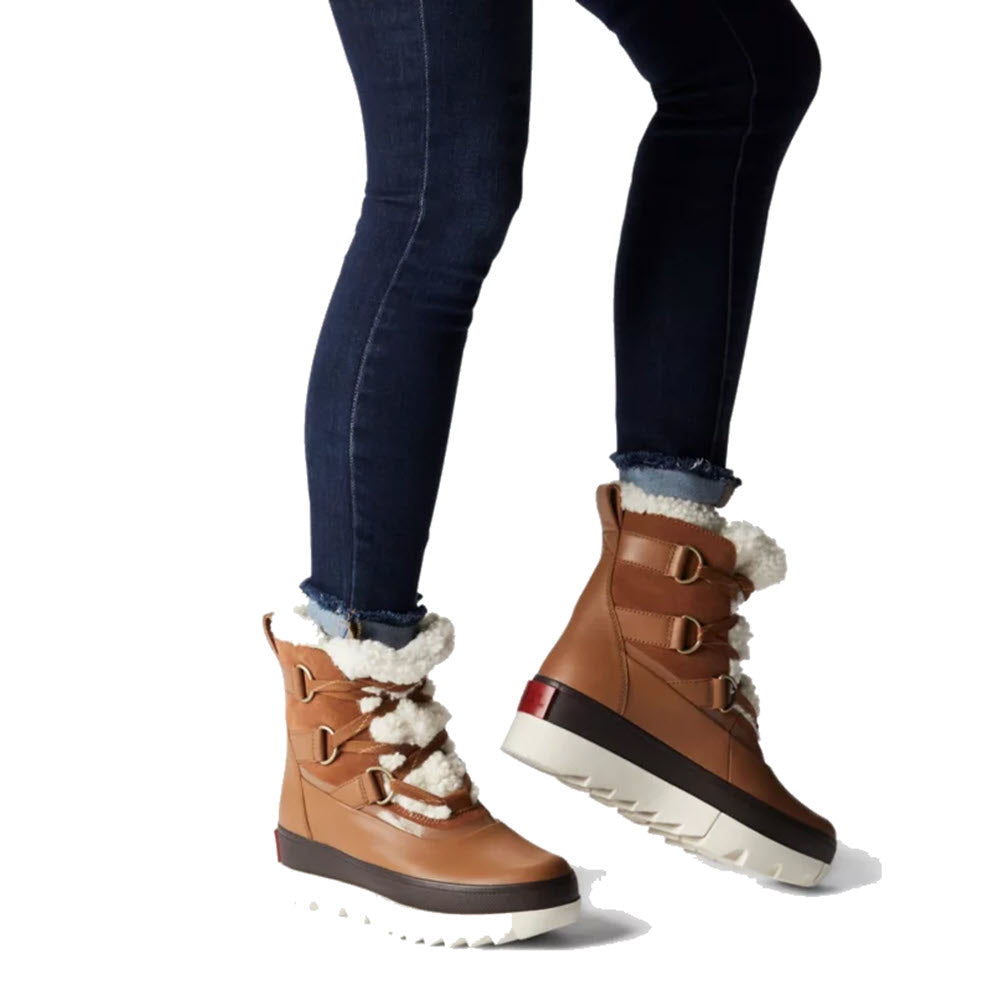 A person wearing Sorel Joan of Arctic Next Boot Velvet Tan - Womens with white fuzzy lining and dark blue jeans on a white background.