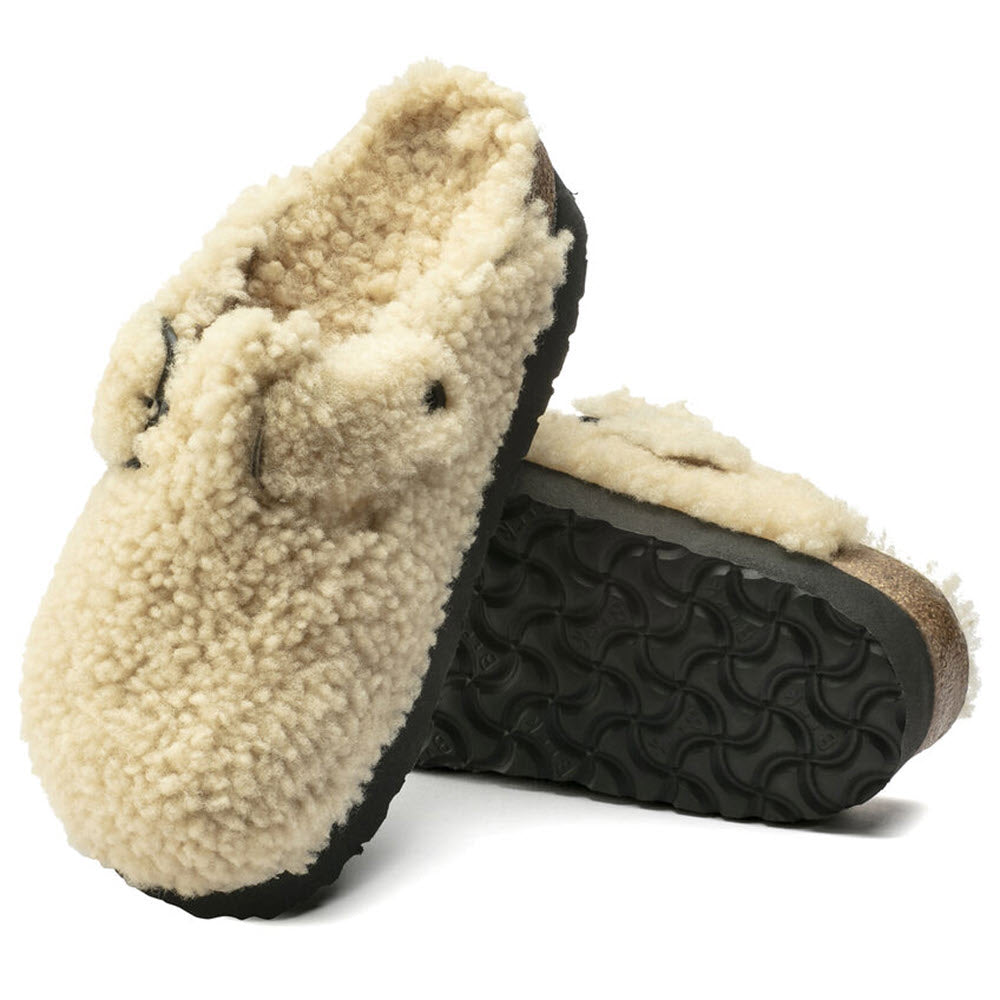 A pair of Birkenstock cream-colored genuine shearling slippers with a black rubber sole, one standing upright and the other lying on its side.