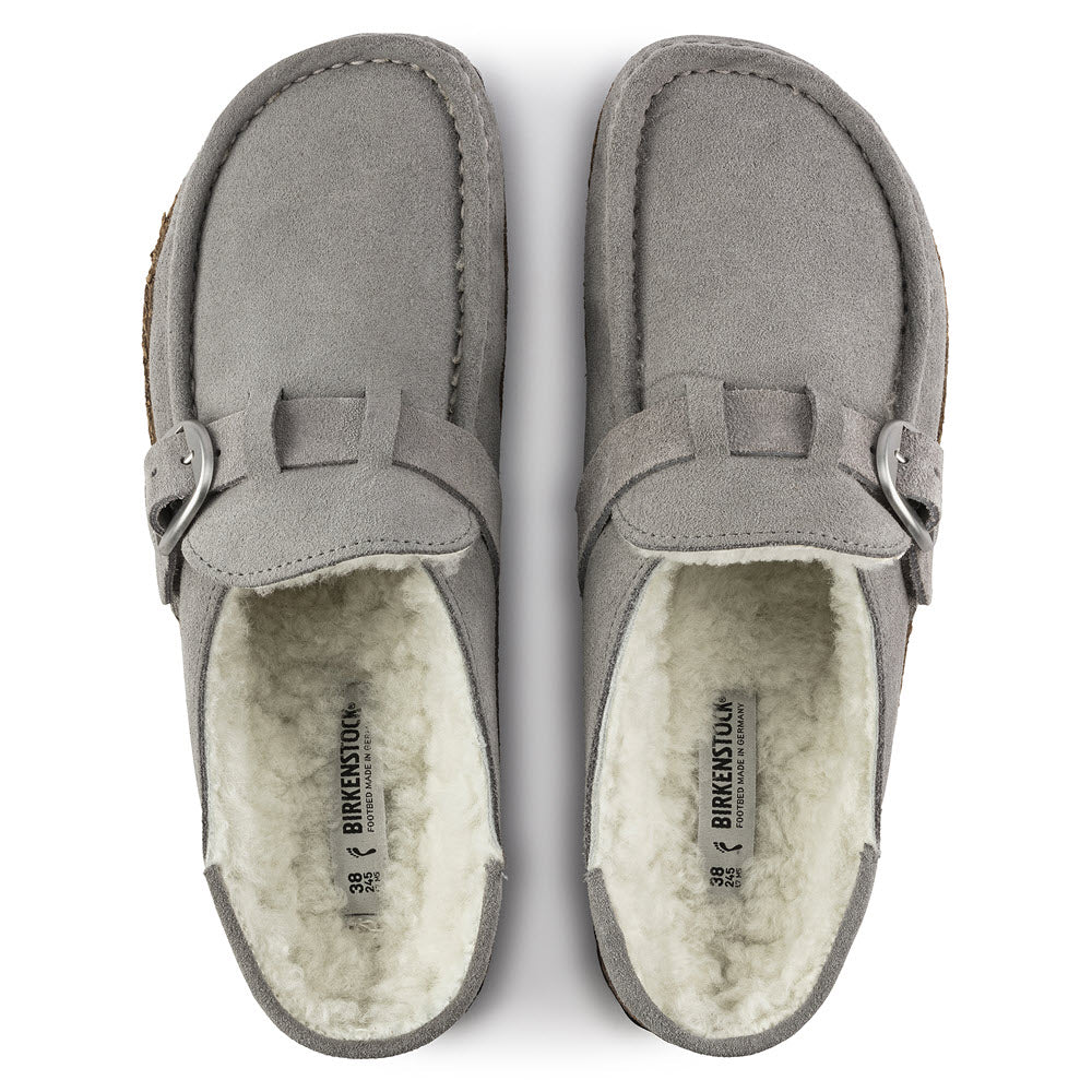 A pair of gray suede leather slippers with white wool lining, displayed with soles facing up, featuring a cork footbed, embodying the Birkenstock Buckley Shearling Stone Coin style.