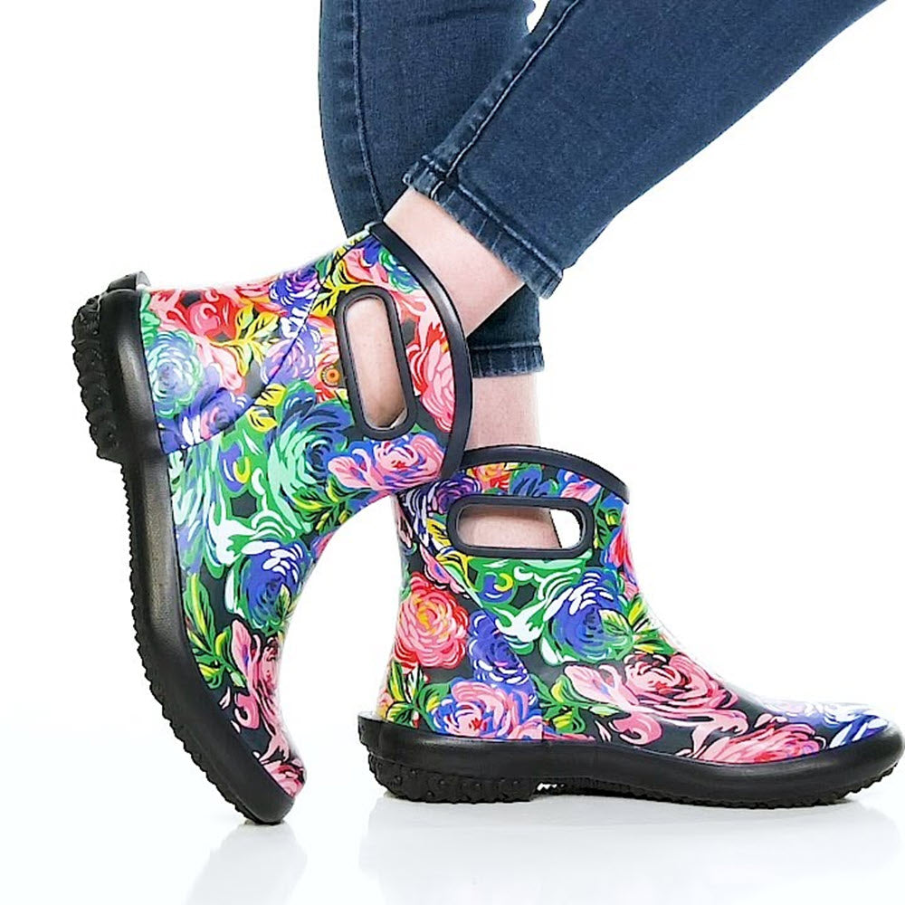 A person wearing BOGS PATCH ANKLE ROSE GARDEN MULTI - WOMENS ankle rain boots with BLOOM eco-friendly footbed, paired with blue jeans, against a white background.