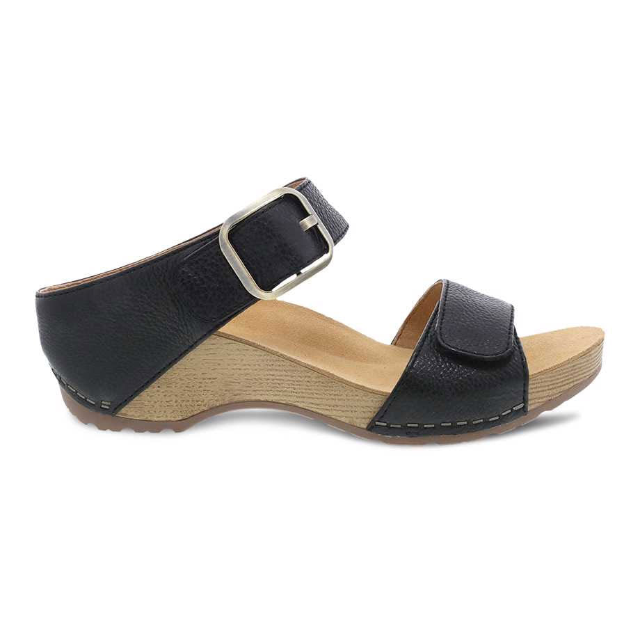 A Dansko Tanya Black burnished leather women&#39;s sandal with a buckle and a low, wooden heel on a white background.