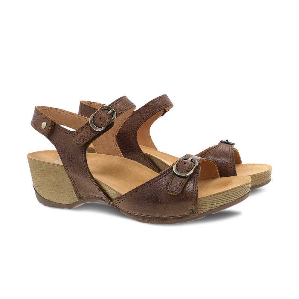 A pair of Dansko Tricia Brown leather wedge sandals with buckle closures, set against a white background.