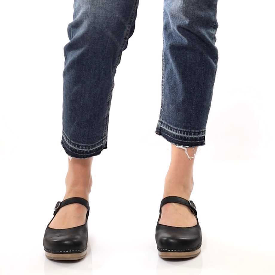 A person wearing distressed, cropped blue jeans and Dansko Bria Black Nubuck mules stands against a white background.