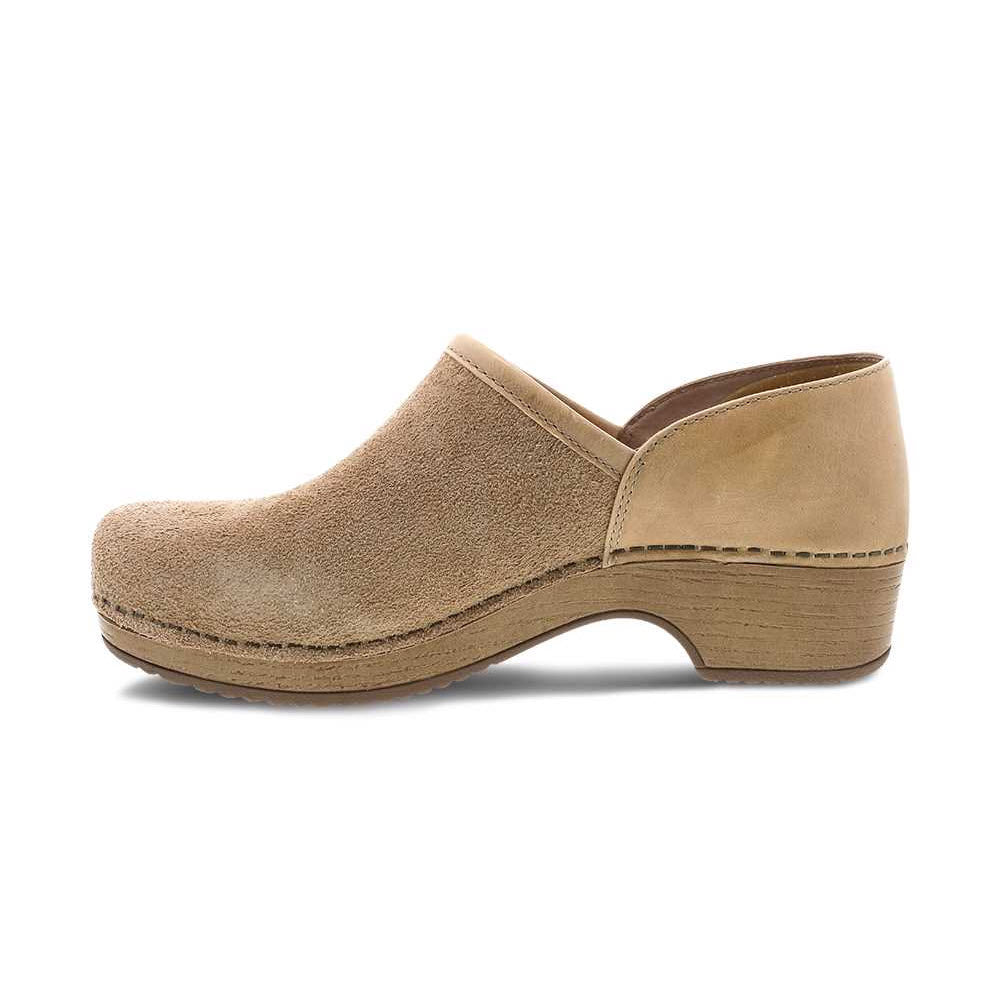 A side view of a single Dansko Brenna Sand Suede slip-on clog with a wooden sole and slight heel, isolated on a white background.