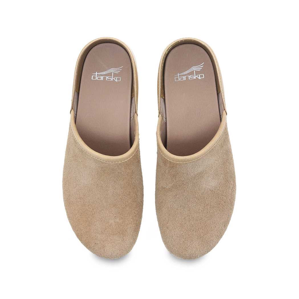 A pair of beige Dansko Brenna Sand Suede clogs with stapled construction viewed from above on a white background.