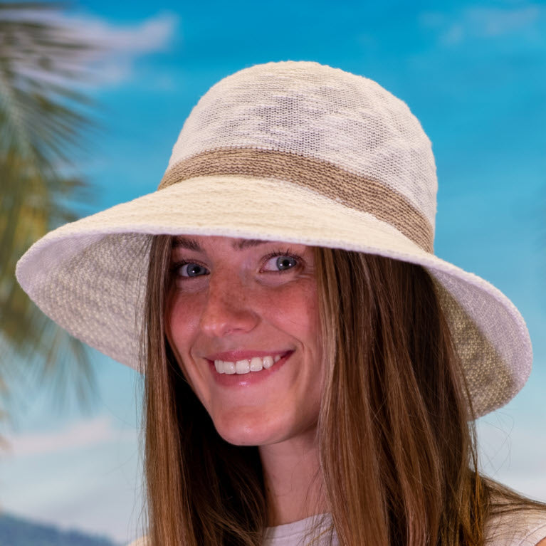 A woman with long brown hair wearing a Shihreen Brim Hat with Stripe Cream smiles in front of a tropical beach backdrop with a palm tree.