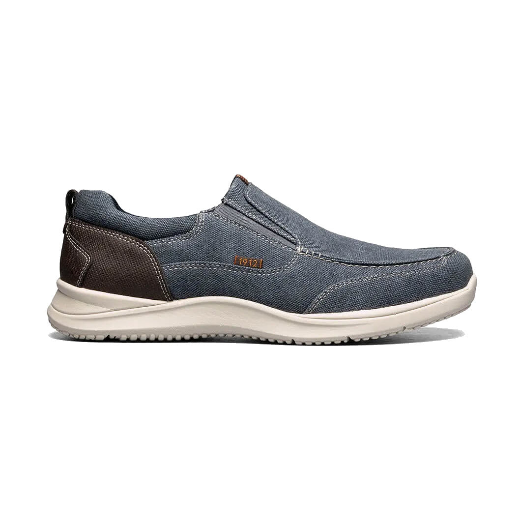 Casual men's Nunn Bush slip-on shoe with blue denim upper and brown leather accents on a white sole, featuring a Memory Foam insole.