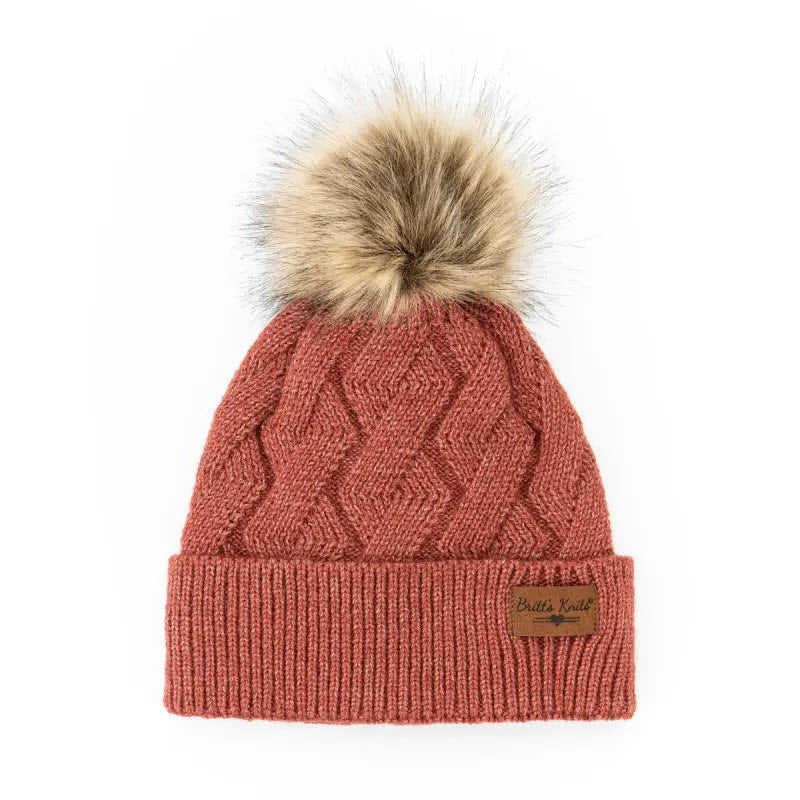 A red BRITTS KNITS POM HAT CEDAR with a fluffy pompom on top and a leather Britts Knits tag on the folded cuff, isolated on a white background.