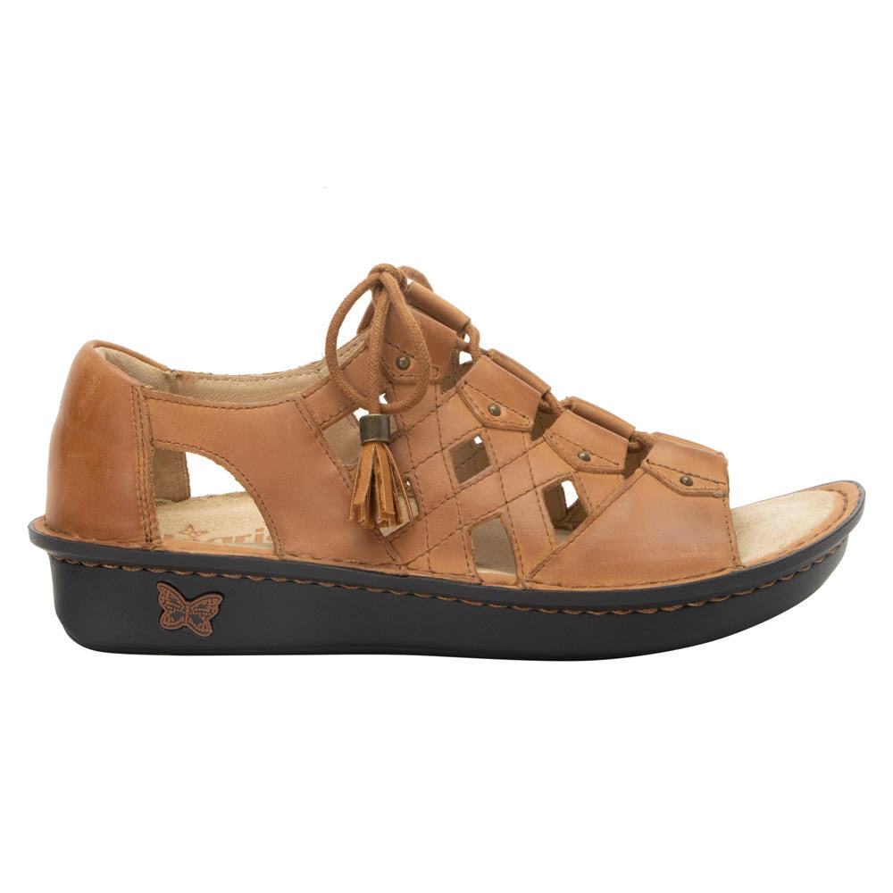Alegria Valerie Cognac gladiator sandals with tassel, metal studs, and a butterfly emblem on a wedge sole, isolated on a white background.