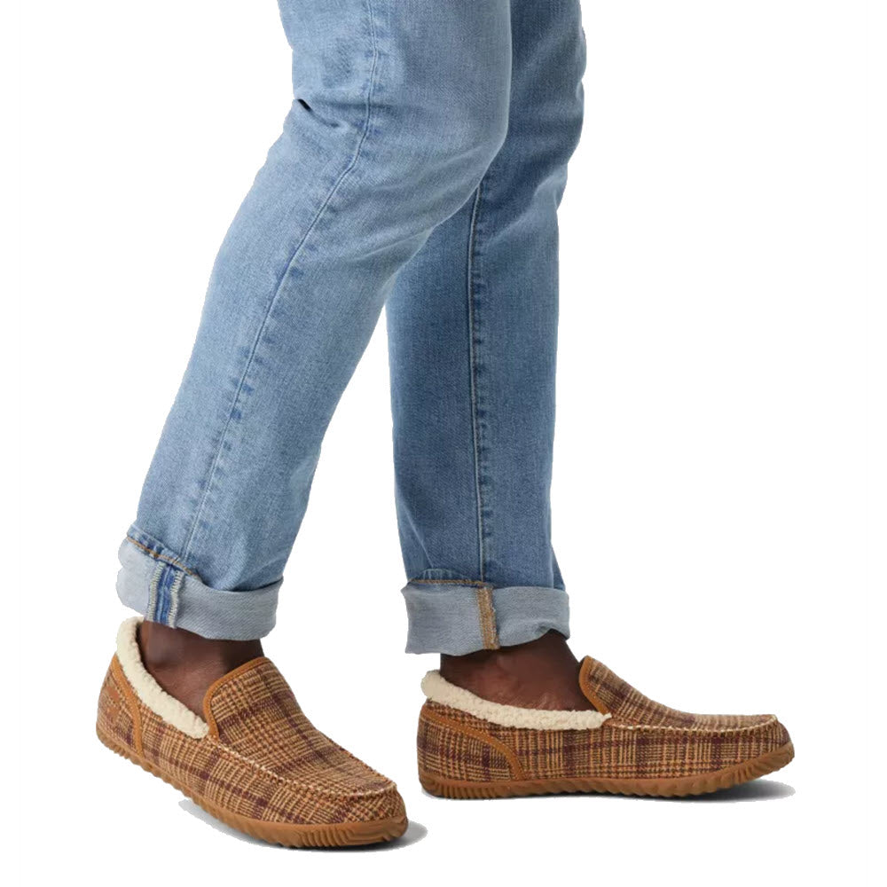 A person wearing blue jeans and brown woven loafers with an EVA footbed, standing against a white background. Only the lower legs and feet are shown wearing Sorel DUDE MOC ELK GUM - MENS.