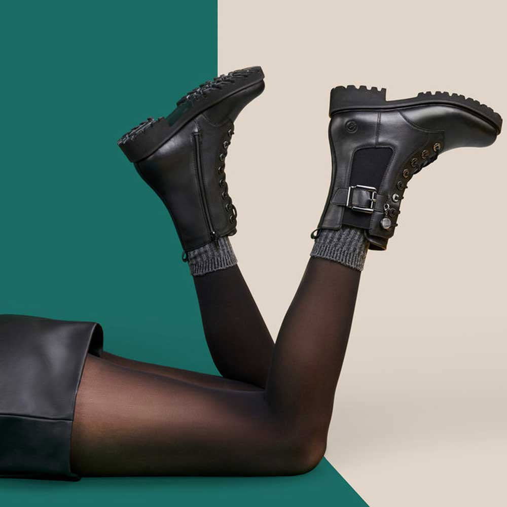 A pair of legs wearing Remonte Tailored Combat Bootie with Buckle Black boots with adjustable laces, elevated against a two-tone teal and green background.