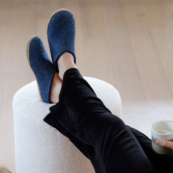 A person lounging with feet up on an ottoman, wearing Glerups The Slip On Leather Denim - Adults slippers and black pants, holding a cup.