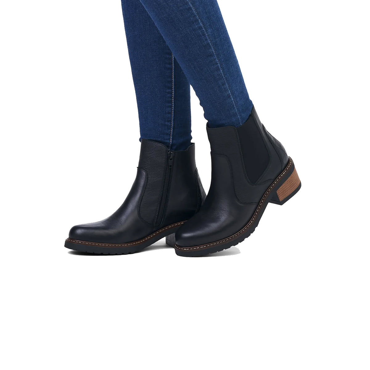 A person wearing dark blue jeans and Remonte Mixed Media Chelsea Bootie Black ankle boots, standing against a white background.