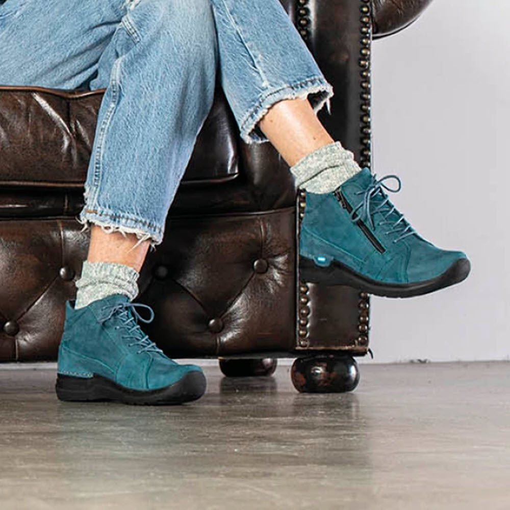 A person wearing Wolky Why Antique Nubuck Petrol ankle-high lace-up shoes and rolled-up jeans sits on a brown leather armchair.